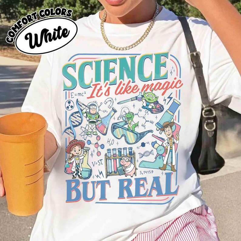 Pu&ograve; includere: T-shirt bianca con grafica colorata con la parola "SCIENCE" e la frase "It's like magic BUT REAL". Il design include personaggi dei cartoni animati e illustrazioni scientifiche. La maglietta ha anche le parole "COMFORT COLORS" e "White".