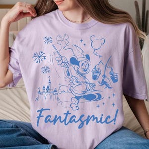 Puede incluir: Una camiseta lila con una ilustraci&oacute;n azul de Mickey Mouse como hechicero, con fuegos artificiales, un castillo y la palabra "Fantasmic!". El dise&ntilde;o es un gr&aacute;fico caprichoso de estilo caricatura.