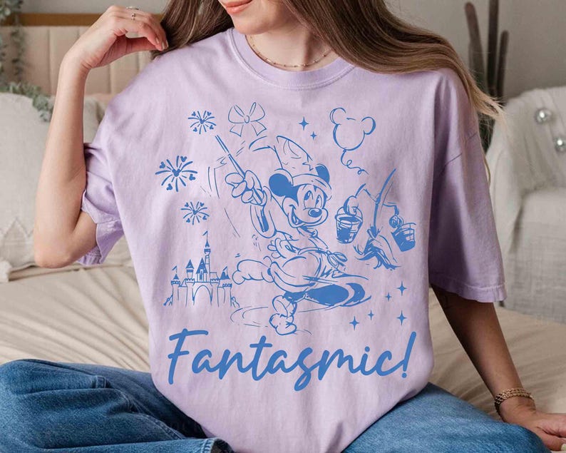 Puede incluir: Una camiseta lila con una ilustraci&oacute;n azul de Mickey Mouse como hechicero, con fuegos artificiales, un castillo y la palabra "Fantasmic!". El dise&ntilde;o es un gr&aacute;fico caprichoso de estilo caricatura.