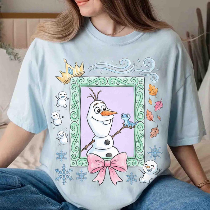 May include: A light blue t-shirt featuring a graphic of Olaf the snowman and a small blue lizard inside a decorative green frame. The design includes a gold crown, a large pink bow, small snowmen, snowflakes, and autumn leaves surrounding the frame.