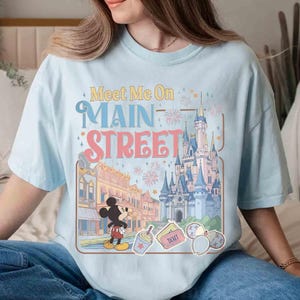 May include: Light blue t-shirt with the text "Meet Me On Main Street" and a graphic featuring a cartoon of Mickey Mouse, a castle, and other theme park elements. The shirt is a relaxed fit.