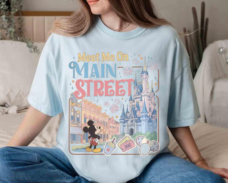 May include: Light blue t-shirt with the text "Meet Me On Main Street" and a graphic featuring a cartoon of Mickey Mouse, a castle, and other theme park elements. The shirt is a relaxed fit.
