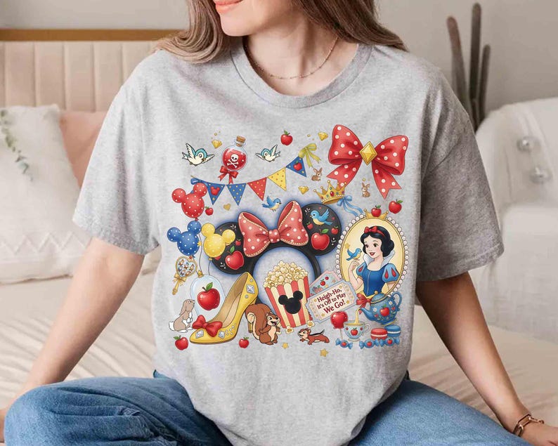 May include: A gray t-shirt featuring a colorful graphic design with Disney-themed elements. The design includes a red polka-dot bow, Snow White, Mickey Mouse ears, popcorn, and the text "Heigh-Ho, It's Off to Play We Go!"