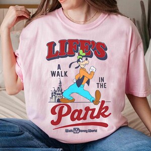 May include: Pink t-shirt featuring a graphic of Goofy walking in front of a castle, with the text "LIFE'S A WALK IN THE PARK" in red and blue. The shirt also includes the Walt Disney World logo.