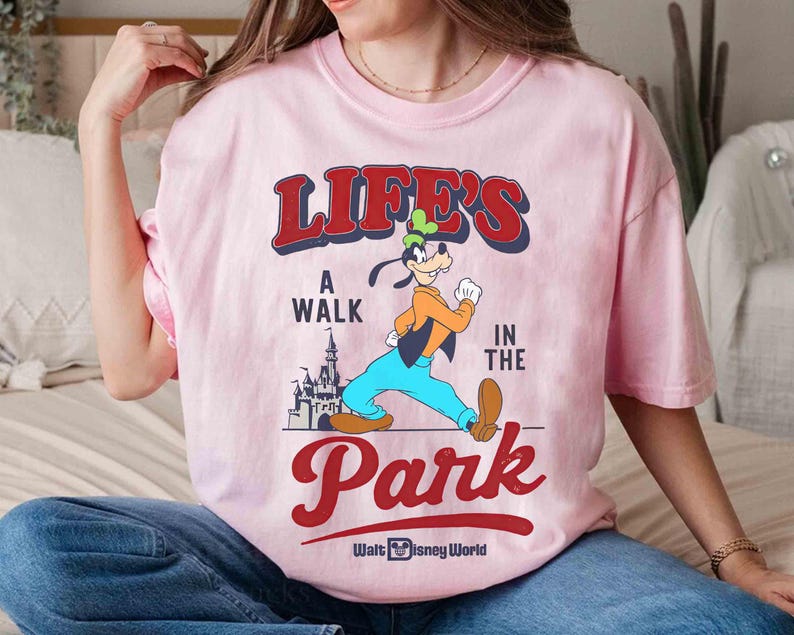 May include: Pink t-shirt featuring a graphic of Goofy walking in front of a castle, with the text "LIFE'S A WALK IN THE PARK" in red and blue. The shirt also includes the Walt Disney World logo.