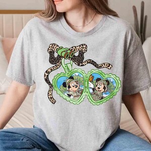 Puede incluir: Una camiseta gris con un estampado de Mickey Mouse y Minnie Mouse con sombreros de safari y sosteniendo binoculares. El estampado est&aacute; rodeado por un dise&ntilde;o de estampado de leopardo.