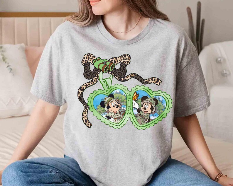 Puede incluir: Una camiseta gris con un estampado de Mickey Mouse y Minnie Mouse con sombreros de safari y sosteniendo binoculares. El estampado est&aacute; rodeado por un dise&ntilde;o de estampado de leopardo.