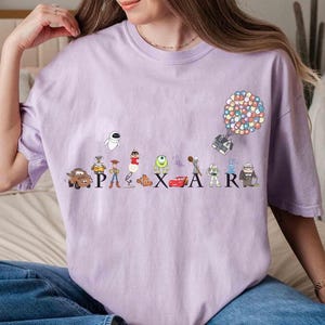 K&ouml;nnte beinhalten: Helllila T-Shirt mit dem Wort "PIXAR" und Zeichentrickfiguren. Das Design zeigt ein Haus, das von einer Gruppe bunter Ballons und anderen Figuren aus beliebten Filmen getragen wird.