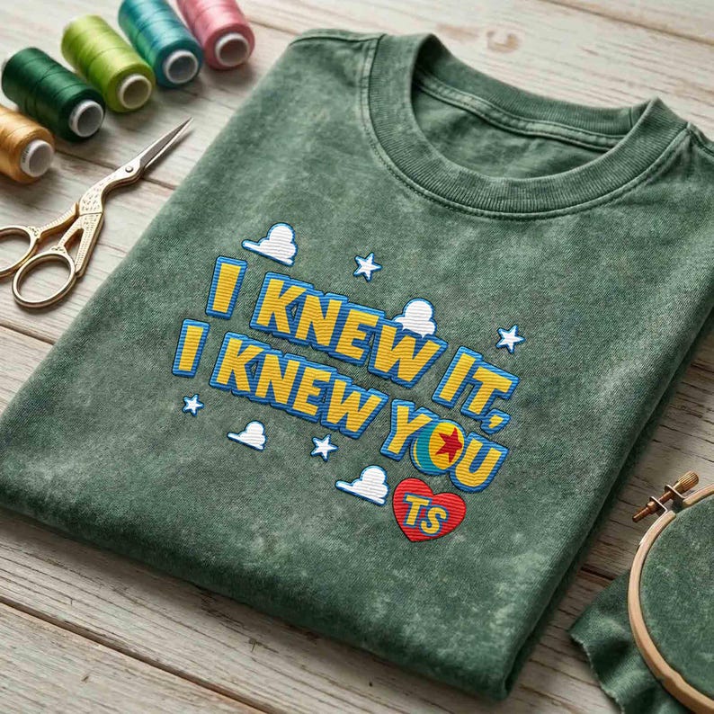 May include: A mineral-washed olive green t-shirt featuring embroidered text that reads I KNEW IT, I KNEW YOU with a red heart containing the letters TS, surrounded by small white embroidered clouds and stars, with sewing supplies nearby.