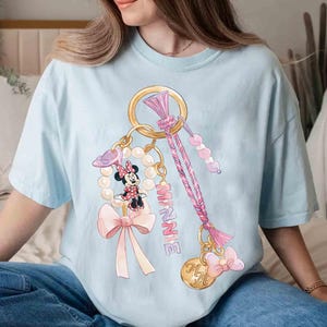 Op de afbeelding: Lichtblauw T-shirt met een Minnie Mouse-ontwerp. De afbeelding toont een gouden sleutelhanger met een parelketting, een Minnie Mouse-figuur, een roze strik en het woord "MINNIE" in roze. Het ontwerp bevat ook een roze en paars gevlochten koord.