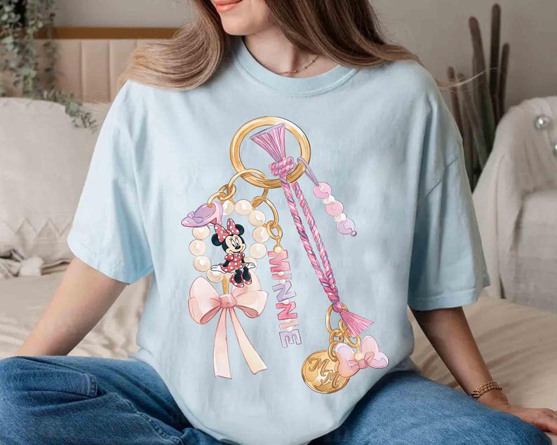 Op de afbeelding: Lichtblauw T-shirt met een Minnie Mouse-ontwerp. De afbeelding toont een gouden sleutelhanger met een parelketting, een Minnie Mouse-figuur, een roze strik en het woord "MINNIE" in roze. Het ontwerp bevat ook een roze en paars gevlochten koord.