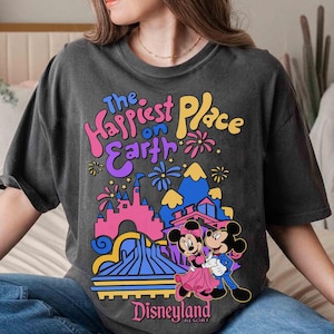 K&ouml;nnte beinhalten: Dunkelgraues T-Shirt mit einem farbenfrohen Disneyland-Motiv mit Mickey und Minnie Mouse. Der Text "The Happiest Place on Earth" ist &uuml;ber der Parkillustration zu sehen. Das Wort "Disneyland" ist unten aufgedruckt.