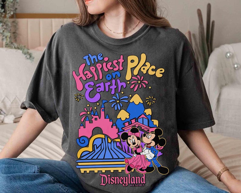 K&ouml;nnte beinhalten: Dunkelgraues T-Shirt mit einem farbenfrohen Disneyland-Motiv mit Mickey und Minnie Mouse. Der Text "The Happiest Place on Earth" ist &uuml;ber der Parkillustration zu sehen. Das Wort "Disneyland" ist unten aufgedruckt.