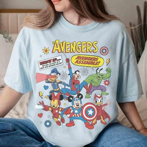 May include: Pale blue t-shirt with a comic-style graphic of Avengers characters. The design includes the word "AVENGERS" and "AVENGERS ASSEMBLE!" alongside illustrations of superheroes. The shirt is a relaxed fit, suitable for everyday wear.