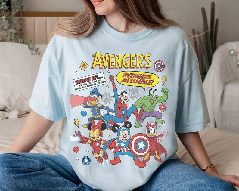 May include: Pale blue t-shirt with a comic-style graphic of Avengers characters. The design includes the word "AVENGERS" and "AVENGERS ASSEMBLE!" alongside illustrations of superheroes. The shirt is a relaxed fit, suitable for everyday wear.