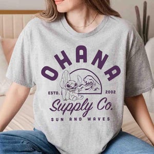 May include: A light gray t-shirt featuring the word "OHANA" in purple block letters above a graphic of a cartoon character with a surfboard and a wave. The text "Supply Co. SUN AND WAVES" is below the graphic.