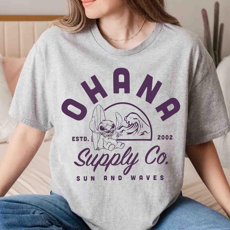 May include: A light gray t-shirt featuring the word "OHANA" in purple block letters above a graphic of a cartoon character with a surfboard and a wave. The text "Supply Co. SUN AND WAVES" is below the graphic.