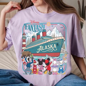 May include: A light purple t-shirt featuring a graphic of a cruise ship with the word "Alaska" and the text "Disney Fantasy". The graphic includes cartoon characters and a lighthouse. The shirt is a comfortable, casual style.