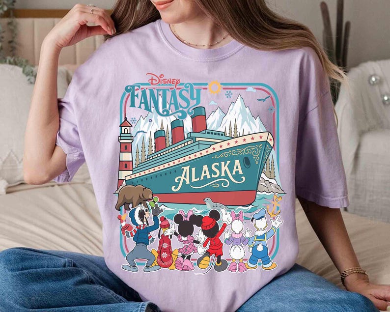 May include: A light purple t-shirt featuring a graphic of a cruise ship with the word "Alaska" and the text "Disney Fantasy". The graphic includes cartoon characters and a lighthouse. The shirt is a comfortable, casual style.