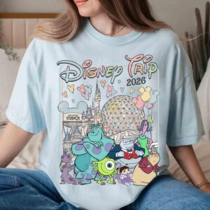 May include: Light blue t-shirt with a colorful graphic of Disney characters and the text "Disney Trip 2026". The graphic includes iconic Disney park landmarks and characters like Sully, Mike Wazowski, and Boo.