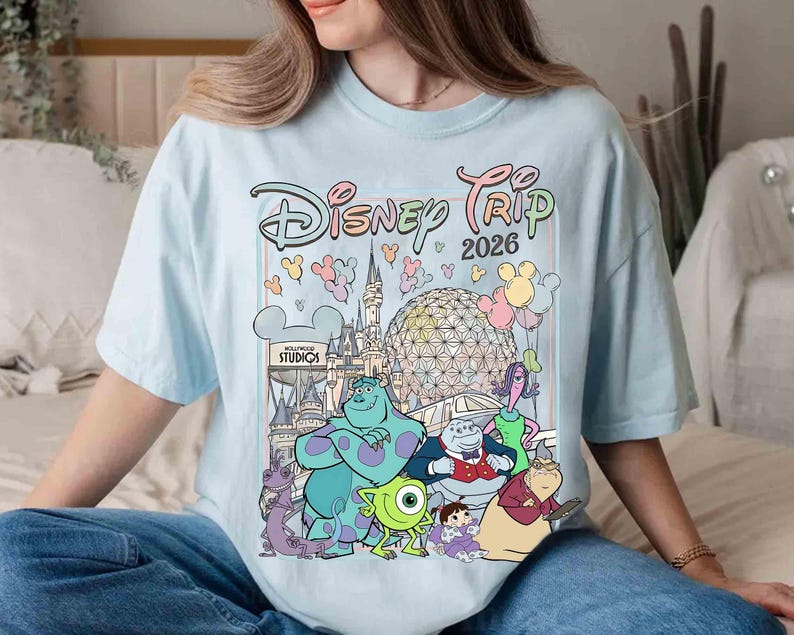 May include: Light blue t-shirt with a colorful graphic of Disney characters and the text "Disney Trip 2026". The graphic includes iconic Disney park landmarks and characters like Sully, Mike Wazowski, and Boo.