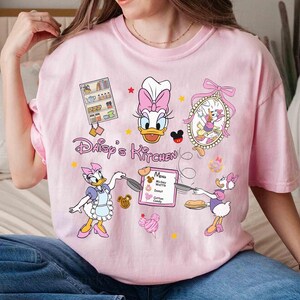 May include: Pink t-shirt with a cartoon image of Daisy Duck in a chef's hat and apron. The shirt says "Daisy's Kitchen" and features other cartoon characters and a menu with items like Mickey Waffles, Donuts, and Cotton Candy.