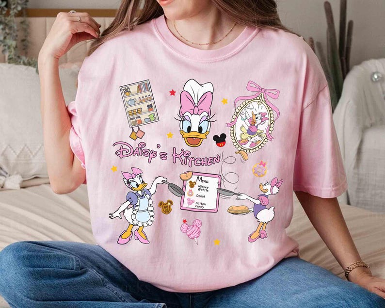 May include: Pink t-shirt with a cartoon image of Daisy Duck in a chef's hat and apron. The shirt says "Daisy's Kitchen" and features other cartoon characters and a menu with items like Mickey Waffles, Donuts, and Cotton Candy.
