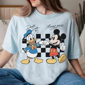 May include: Light blue t-shirt featuring Donald Duck and Mickey Mouse. Donald Duck is shown fist-bumping Mickey Mouse in front of a black and white checkered background. The text "Donald Duck #1" and "Mickey Mouse" are above the characters.