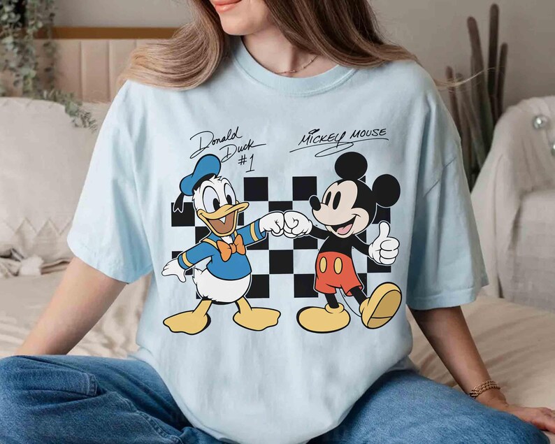 May include: Light blue t-shirt featuring Donald Duck and Mickey Mouse. Donald Duck is shown fist-bumping Mickey Mouse in front of a black and white checkered background. The text "Donald Duck #1" and "Mickey Mouse" are above the characters.