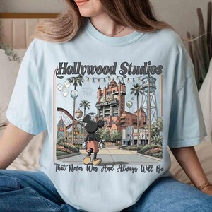 K&ouml;nnte beinhalten: Hellblaues T-Shirt mit einer Grafik von Hollywood Studios und Mickey Mouse. Das Design enth&auml;lt den Text "Hollywood Studios" und "That Never Was And Always Will Be". Das Bild zeigt eine Themenpark-Szene mit Geb&auml;uden und Attraktionen.