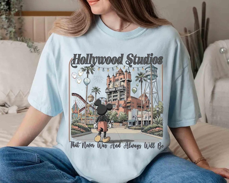 K&ouml;nnte beinhalten: Hellblaues T-Shirt mit einer Grafik von Hollywood Studios und Mickey Mouse. Das Design enth&auml;lt den Text "Hollywood Studios" und "That Never Was And Always Will Be". Das Bild zeigt eine Themenpark-Szene mit Geb&auml;uden und Attraktionen.
