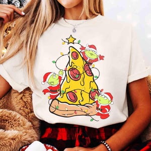 Op de afbeelding: Cr&egrave;mekleurig T-shirt met een pizzapunt versierd als kerstboom. De pizza heeft pepperoni en is versierd met lichtslingers en een ster. Drie groene alien-personages in kerstmutsen en -pakken zijn ook afgebeeld.