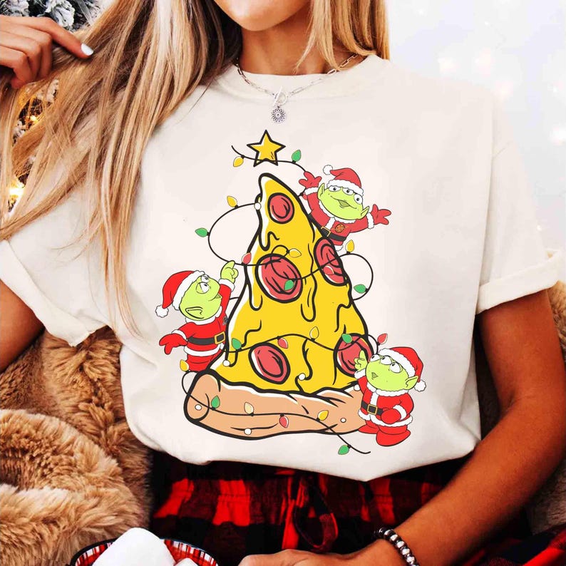 Op de afbeelding: Cr&egrave;mekleurig T-shirt met een pizzapunt versierd als kerstboom. De pizza heeft pepperoni en is versierd met lichtslingers en een ster. Drie groene alien-personages in kerstmutsen en -pakken zijn ook afgebeeld.