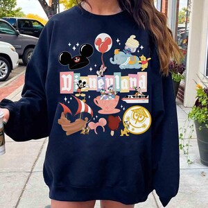 May include: Navy blue sweatshirt featuring a colorful Disneyland graphic. The design includes Mickey Mouse, Minnie Mouse, Dumbo, Winnie the Pooh, and other Disney characters. The word "Disneyland" is prominently displayed in a playful font.
