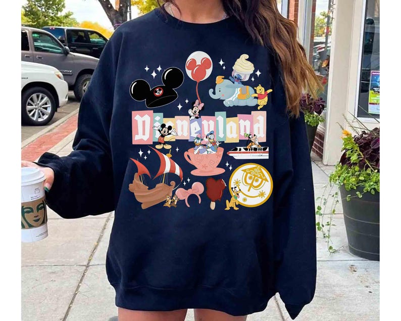 May include: Navy blue sweatshirt featuring a colorful Disneyland graphic. The design includes Mickey Mouse, Minnie Mouse, Dumbo, Winnie the Pooh, and other Disney characters. The word "Disneyland" is prominently displayed in a playful font.