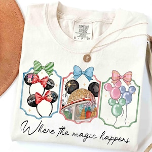 May include: Cream-colored t-shirt featuring three framed illustrations of Mickey Mouse ears, a theme park, and balloons. The text "Where the magic happens" is written in cursive below the illustrations. A gold necklace is draped on the shirt.