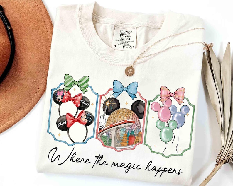 May include: Cream-colored t-shirt featuring three framed illustrations of Mickey Mouse ears, a theme park, and balloons. The text "Where the magic happens" is written in cursive below the illustrations. A gold necklace is draped on the shirt.