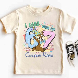 May include: Cream-colored children's t-shirt with a colorful graphic. The graphic says "I WAS 6 NOW I'M 7" with a cartoon chipmunk character. Black canvas sneakers with white laces are next to the shirt.