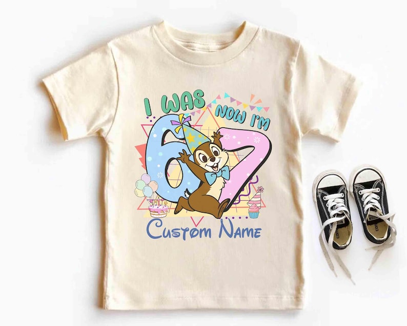 May include: Cream-colored children's t-shirt with a colorful graphic. The graphic says "I WAS 6 NOW I'M 7" with a cartoon chipmunk character. Black canvas sneakers with white laces are next to the shirt.