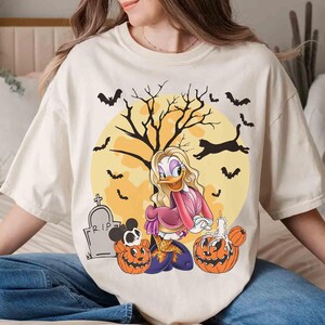 Peut inclure: T-shirt cr&egrave;me avec un motif d'Halloween. Le motif comprend un personnage de dessin anim&eacute;, des citrouilles, des chauves-souris, un arbre et une pleine lune. Le t-shirt a une coupe d&eacute;contract&eacute;e et des manches courtes.