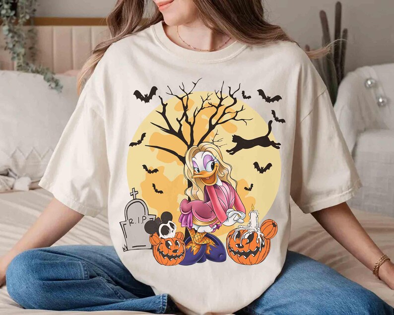 Peut inclure: T-shirt cr&egrave;me avec un motif d'Halloween. Le motif comprend un personnage de dessin anim&eacute;, des citrouilles, des chauves-souris, un arbre et une pleine lune. Le t-shirt a une coupe d&eacute;contract&eacute;e et des manches courtes.