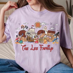 May include: A lilac t-shirt with a pirate-themed design featuring the numbers "25" and cartoon characters. The text "The Lee Family" is printed in red script below. The design includes ships, a sun, and birds, creating a fun, family-oriented theme.
