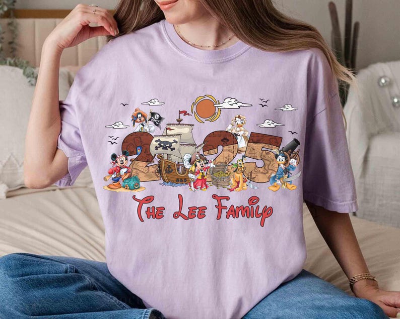 May include: A lilac t-shirt with a pirate-themed design featuring the numbers "25" and cartoon characters. The text "The Lee Family" is printed in red script below. The design includes ships, a sun, and birds, creating a fun, family-oriented theme.
