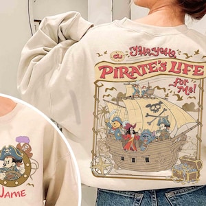 K&ouml;nnte beinhalten: Cremefarbenes Sweatshirt mit einem Piratenschiff-Design mit Disney-Figuren. Auf der R&uuml;ckseite des Sweatshirts steht der Text "Yo Ho, Yo Ho, A Pirate's Life For Me!". Auf der Vorderseite befindet sich ein Mickey Mouse Piraten-Design mit dem Wort "Name".