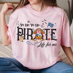 Peut inclure: Un t-shirt rose clair avec un motif sur le th&egrave;me des pirates. Le mot "PIRATE" est affich&eacute; dans une police audacieuse, avec un canard de dessin anim&eacute; portant un chapeau de pirate et tenant une &eacute;p&eacute;e. Les phrases "Yo Ho Yo Ho" et "Life for me" sont &eacute;galement incluses.