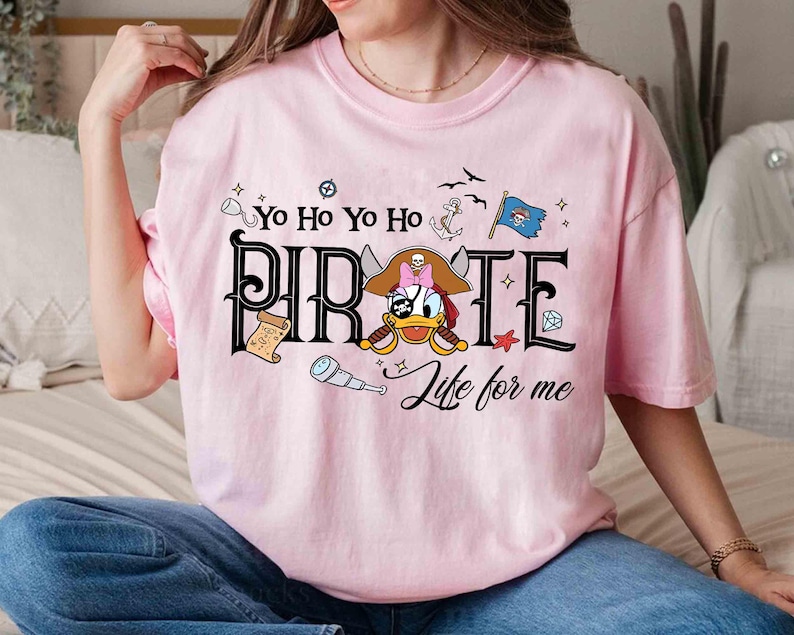 Peut inclure: Un t-shirt rose clair avec un motif sur le th&egrave;me des pirates. Le mot "PIRATE" est affich&eacute; dans une police audacieuse, avec un canard de dessin anim&eacute; portant un chapeau de pirate et tenant une &eacute;p&eacute;e. Les phrases "Yo Ho Yo Ho" et "Life for me" sont &eacute;galement incluses.