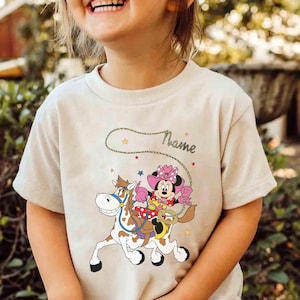 May include: Beige t-shirt featuring a cartoon Minnie Mouse riding a horse, wearing a pink cowgirl hat and holding a lasso. The word "Name" is written above the graphic in a cursive font. Stars and a rope design complete the illustration.