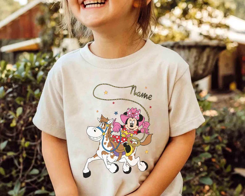 May include: Beige t-shirt featuring a cartoon Minnie Mouse riding a horse, wearing a pink cowgirl hat and holding a lasso. The word "Name" is written above the graphic in a cursive font. Stars and a rope design complete the illustration.