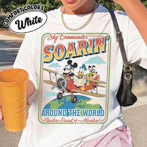 May include: White t-shirt featuring a colorful graphic of Mickey Mouse, Donald Duck, and Goofy in a biplane, with the words "Sky Commander Soarin' Around The World". The shirt also has the words "Comfort Colors" and "White" in a circle.