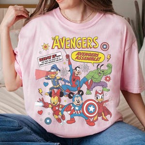May include: A pale pink t-shirt with a comic book style design of Disney characters as Avengers. The graphic includes the words "AVENGERS" and "AVENGERS ASSEMBLE!" and other text. The shirt has a round neck and short sleeves.